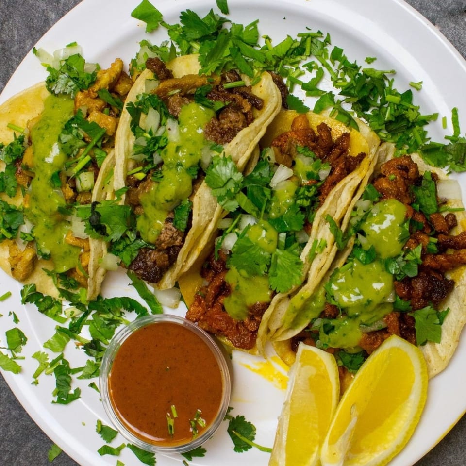 A plate of Golden Ox Brundage tacos with green sauce, cilantro, onions, and lemon wedges.